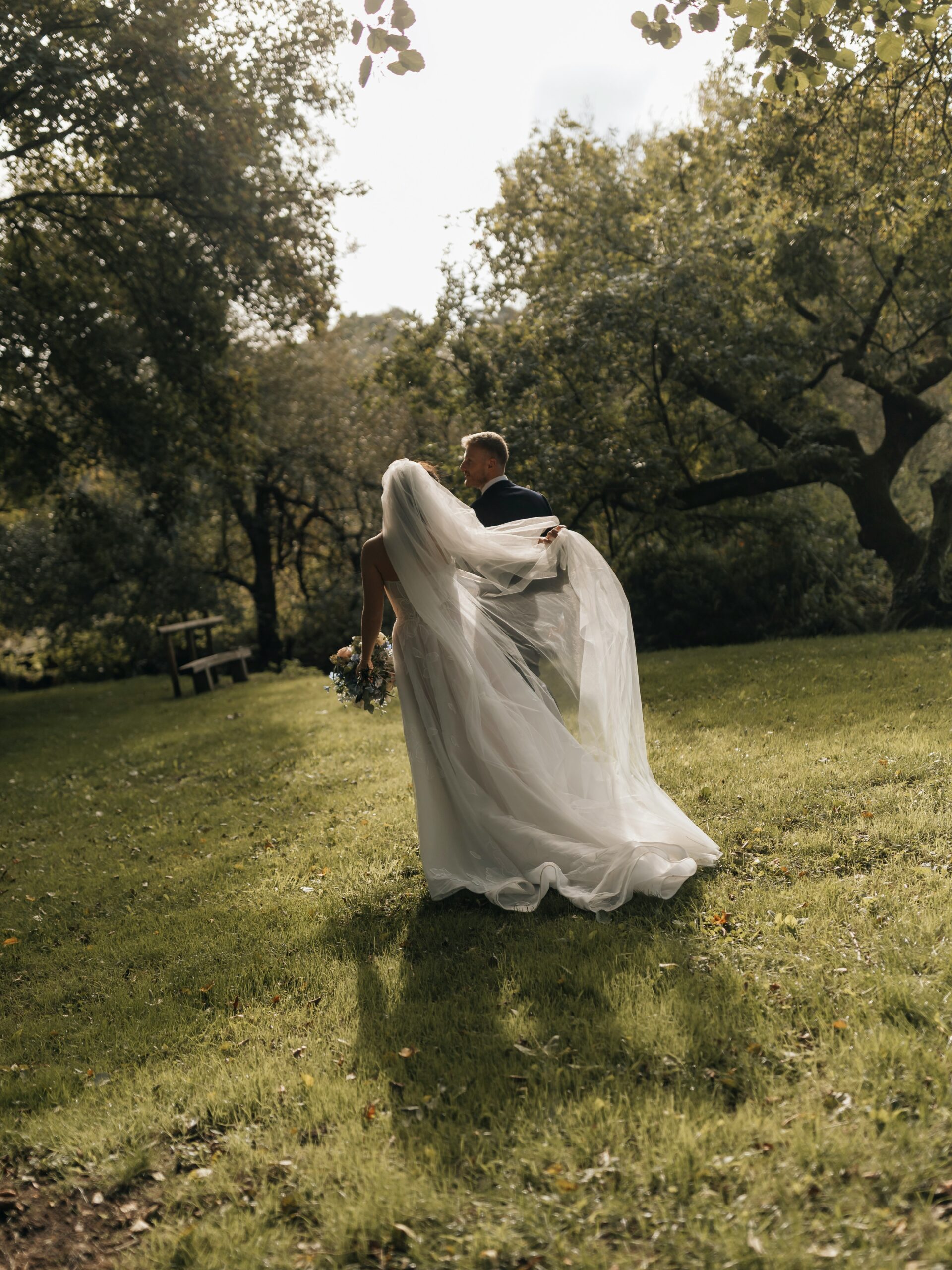 A couple walking among old trees, her veil lifted by the wind